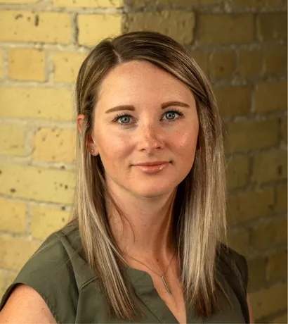Blonde woman with green top smiling at the camera in front of a brick wall.