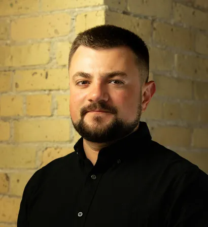 Man with a beard wearing a black shirt, standing in front of a brick wall.